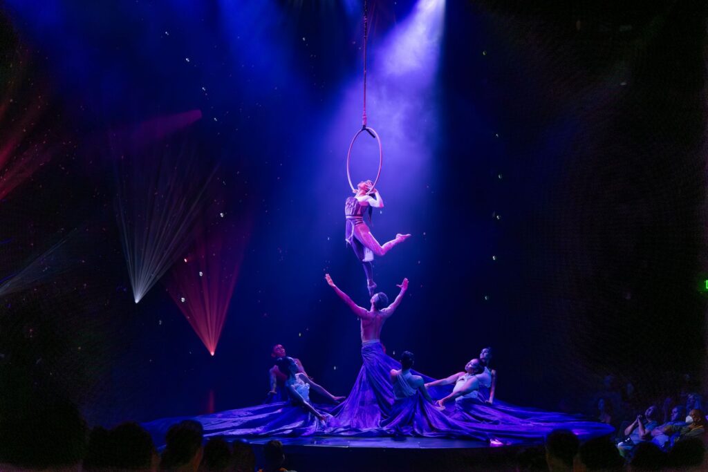 Aerial performer suspended above the stage in a blue spotlight during Auana.