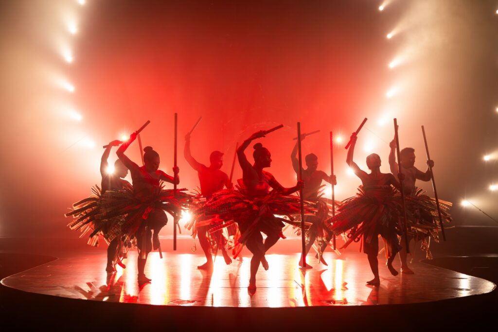 Silhouettes of dancers performing a group routine in bright red-orange lighting during Auana.