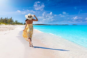 A woman walking on a Bahamian beach wearing a yellow dress and hat next to turquoise water.
