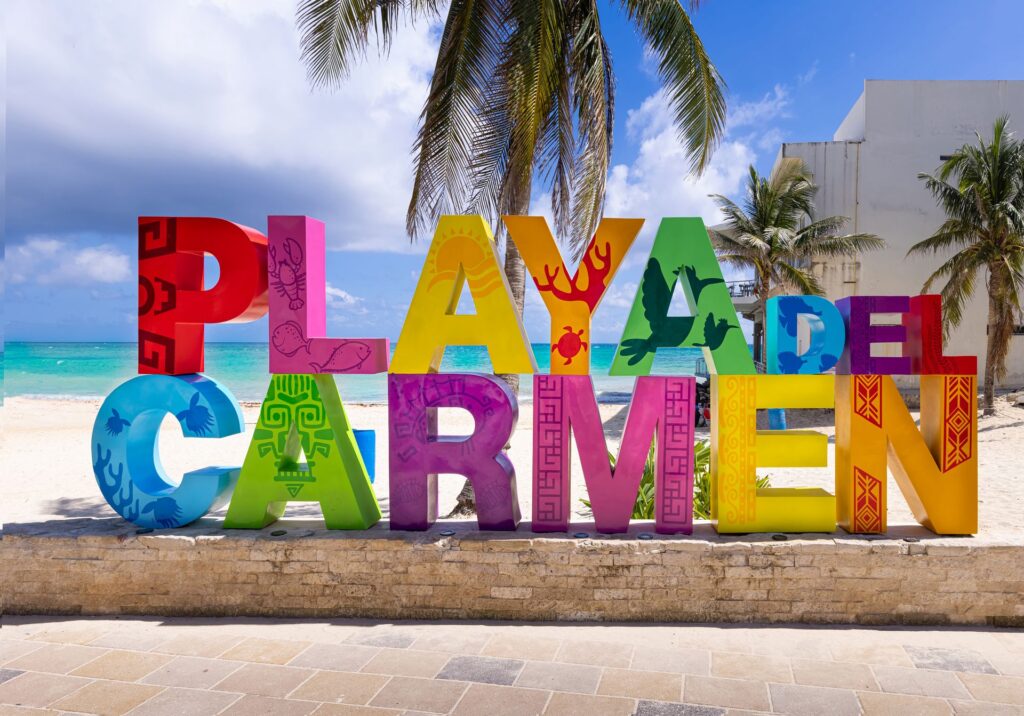 Colorful Playa del Carmen sign near the beach with palm trees and turquoise water in the background.