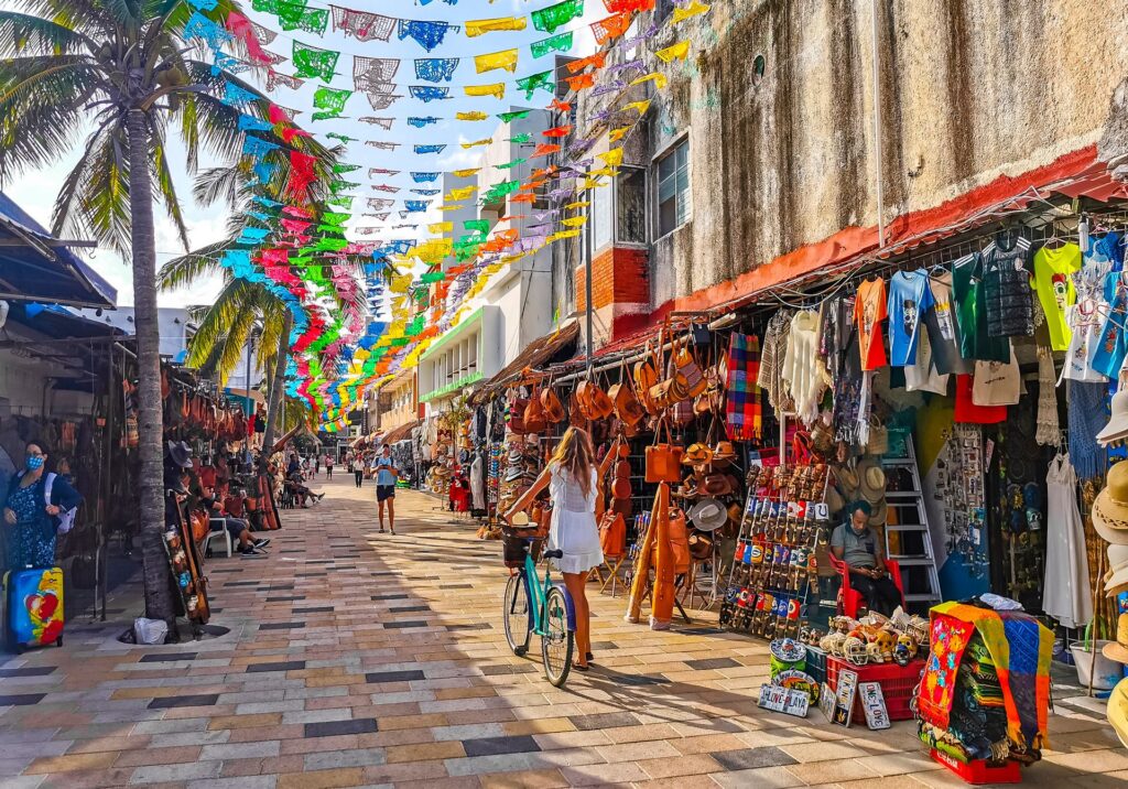 Colorful market street in Playa del Carmen with hanging papel picado banners, palm trees, artisan shops, and a woman walking her bicycle during the day.