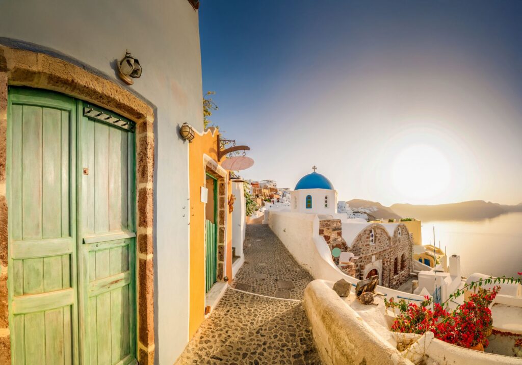 harming cobblestone street in Oia, Santorini at golden hour, featuring pastel houses, traditional doors, and a blue-domed church with sea views.