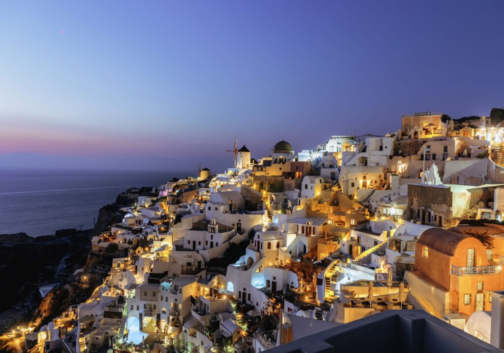 The panoramic view of the old town of Oia or Ia on the island of Santorini, Greece, features, whitewashed houses, blue dome churches, and traditional windmills illuminated with street lights after sunset at blur hours. The picturesque landscape captures the beauty of the picturesque sunset in la Village, a famous spot on the Greek islands of Santorini town. The colorful buildings on the cliff side add to the picturesque atmosphere in Cyclades, Greece.