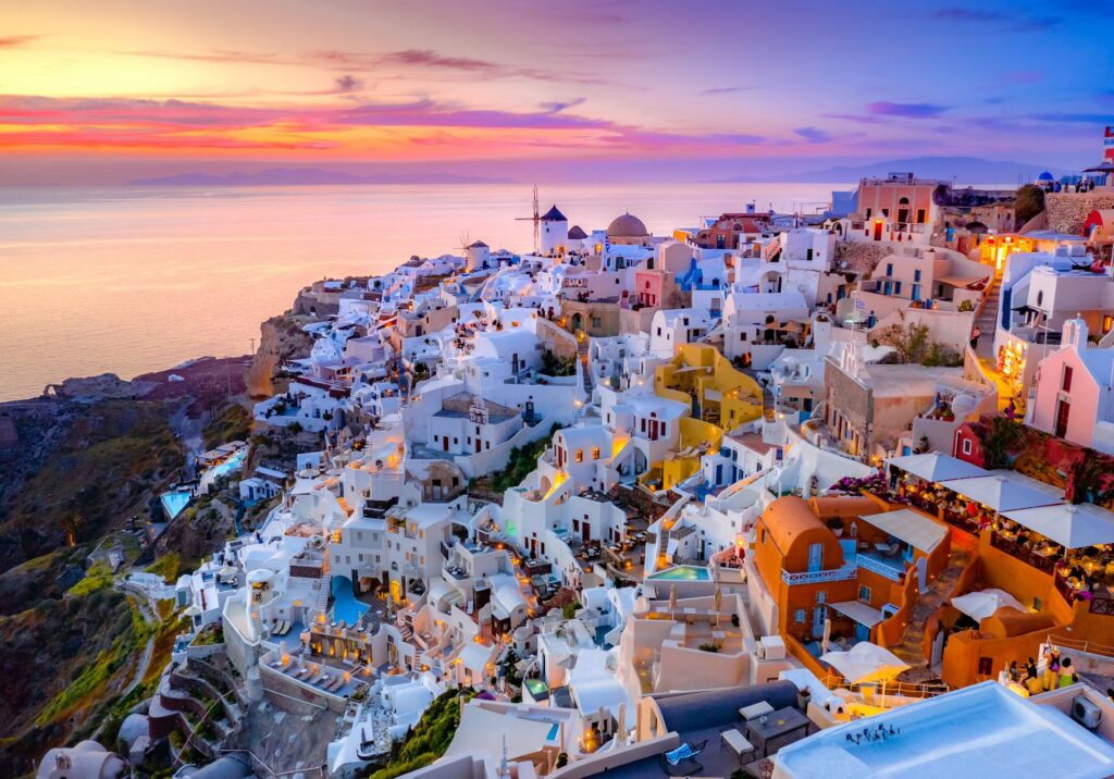 Panoramic sunset view of Oia, Santorini, with whitewashed buildings cascading down the cliffs toward the Aegean Sea. Warm golden light and pastel skies illuminate the island’s iconic architecture.