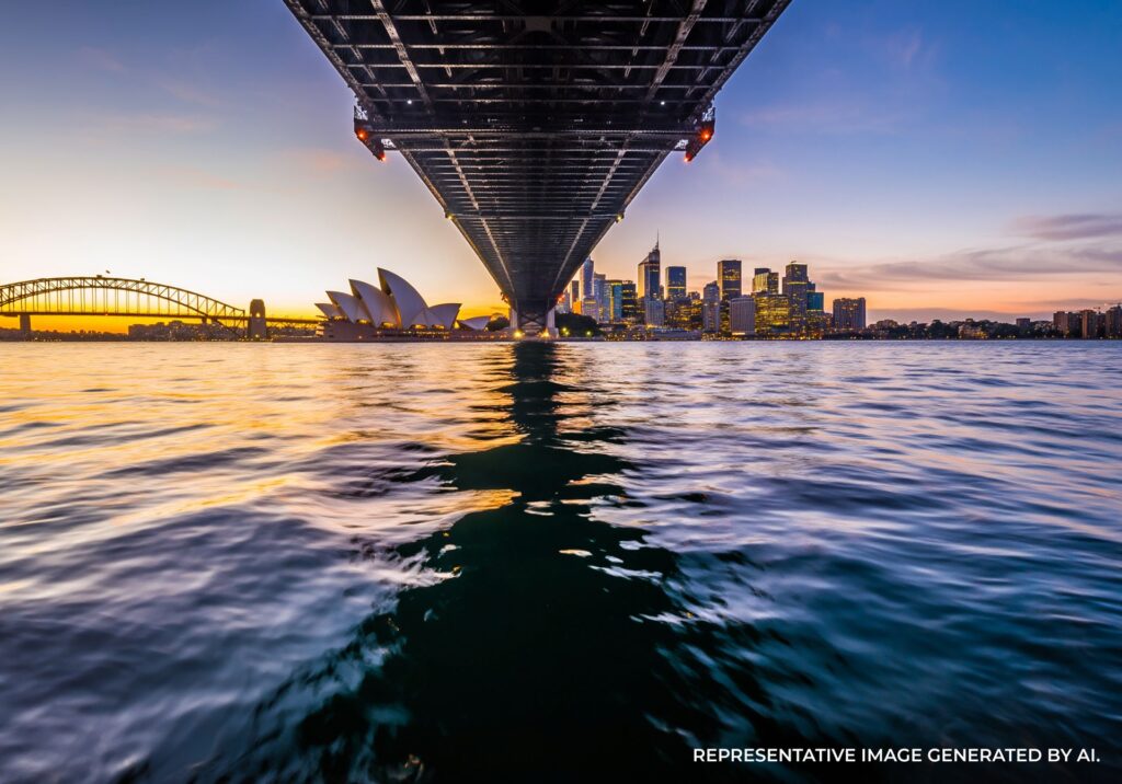 Sunrise glow beneath the Sydney Harbour Bridge with the skyline reflecting gently on calm water.