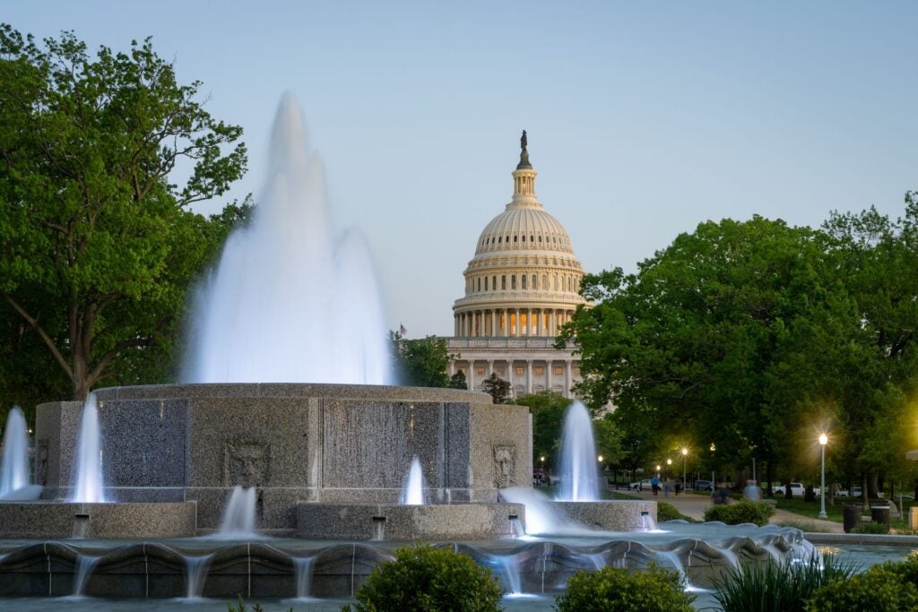 The U.S. Capitol dome behind the Bartholdi Fountain in Washington, D.C., surrounded by trees in the early evening light.