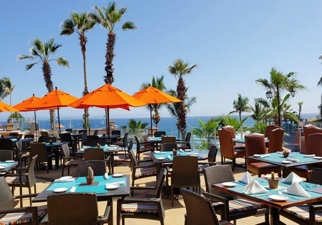 Restaurant patio with orange umbrellas and ocean views surrounded by palm trees at a resort in Cabo San Lucas Mexico