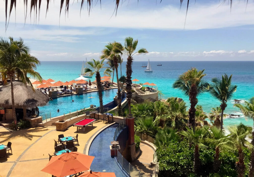 Outdoor restaurant terrace with turquoise tables and orange umbrellas overlooking palm trees and the ocean at a resort in Cabo San Lucas Mexico