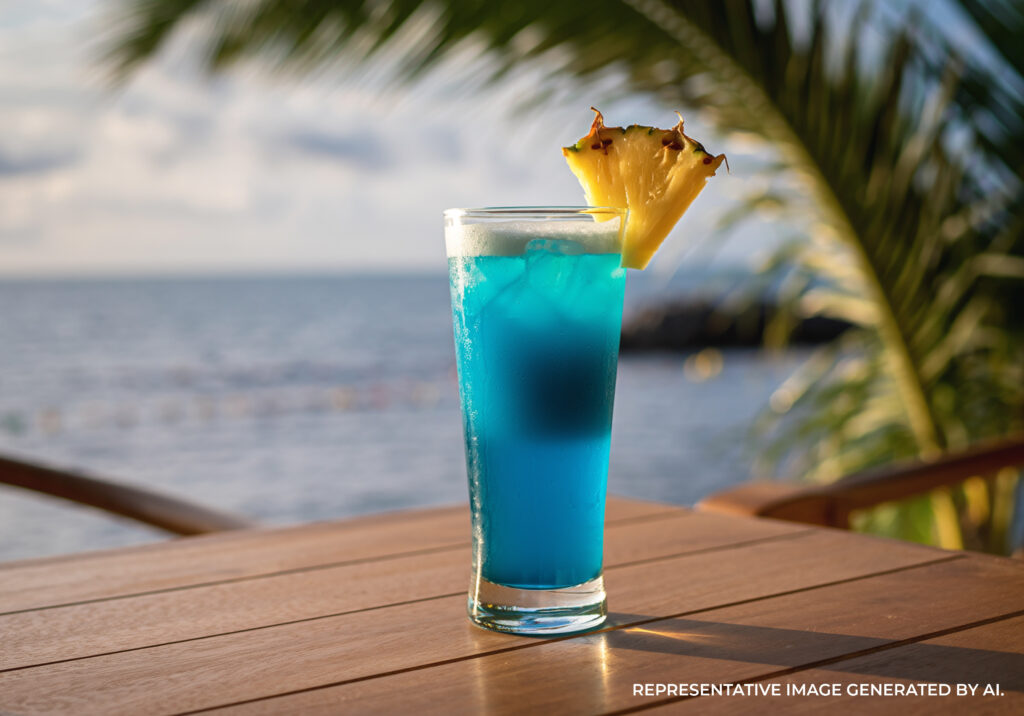 A Blue Hawaiian cocktail garnished with a pineapple wedge on a wooden table overlooking the ocean on the Island of Kaua'i.