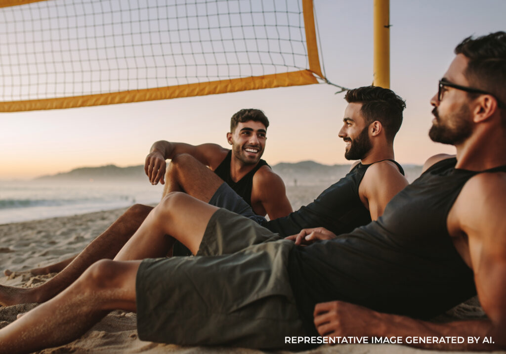 Two men sitting on the sand near a volleyball net at sunset laughing with the shoreline in the background.