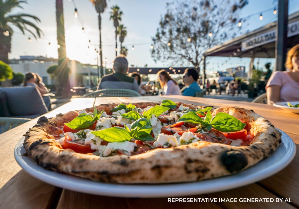 A freshly baked pizza served with drinks on a wooden table near the beach with palm trees.