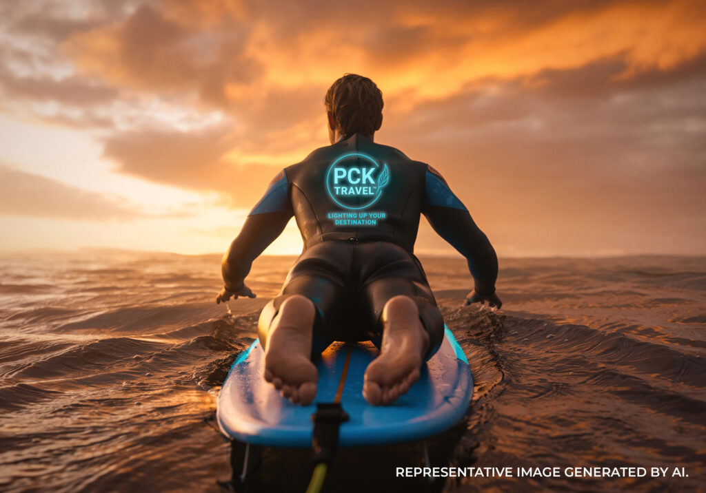 A person wearing a PCK Travel branded wetsuit paddling a surf board across the ocean at sunset.