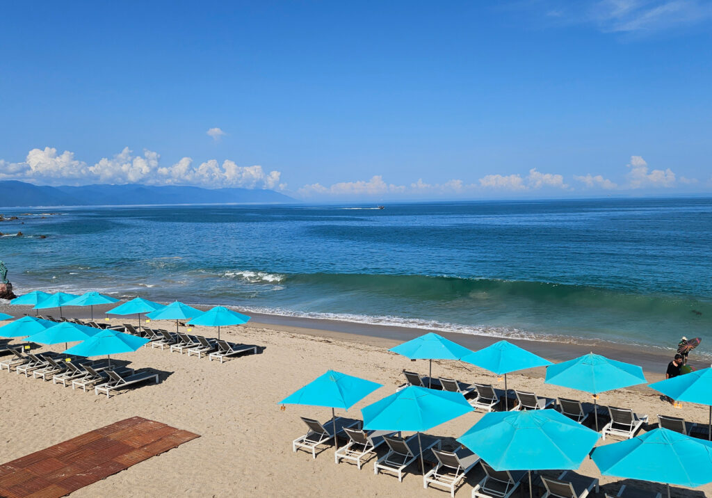 Row of bright turquoise umbrellas and lounge chairs set along a sunny beach in Puerto Vallarta with gentle waves in the background