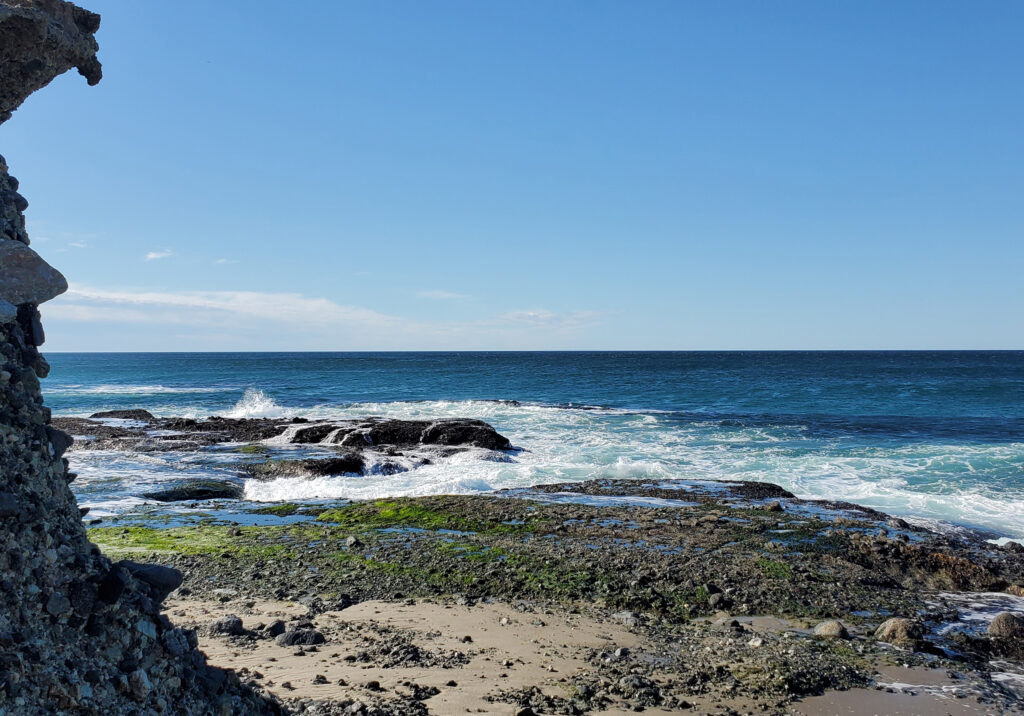 Rocky shoreline at Newport Beach, California with waves crashing against the rocks under a clear blue sky.