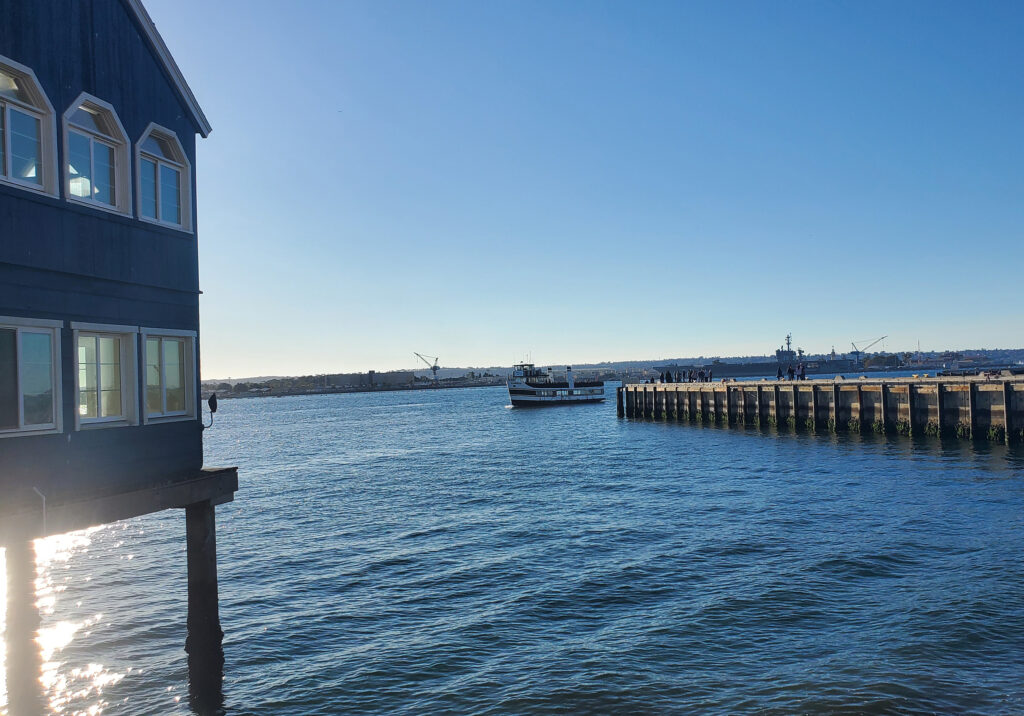 Calm blue waters of San Diego Bay with boats, marina structures, and shoreline under a bright sky.