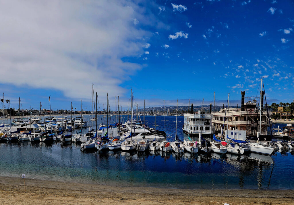 Wide view of Mission Bay Marina with sailboats docked across calm blue water beneath scattered white clouds.