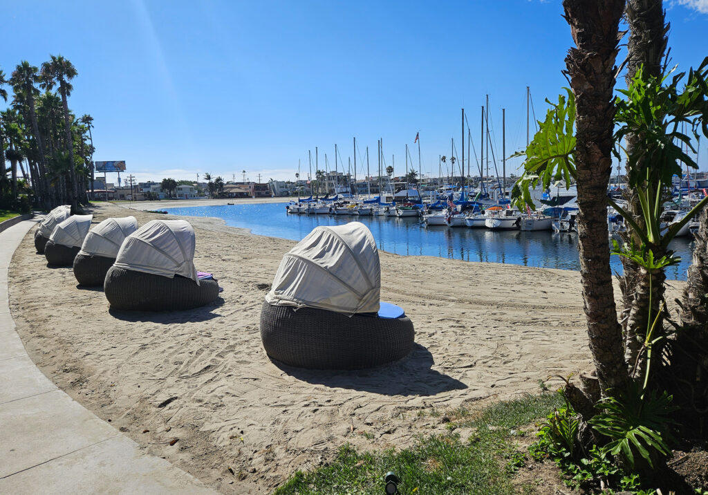 Beach cabanas and stone seating areas along a sandy shoreline with a marina full of sailboats in the background.