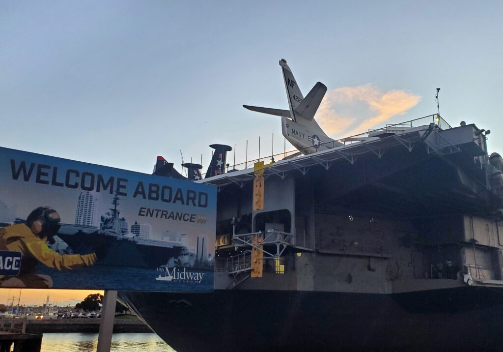 Back view of the USS Midway Museum aircraft carrier with planes visible on deck and harbor waters below.