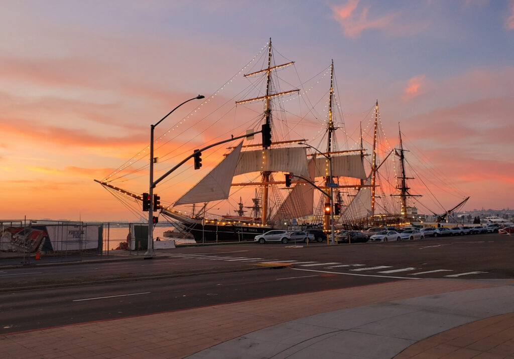 Historic tall ship sailing at sunset in San Diego Harbor, with dramatic golden sky and calm ocean waters.