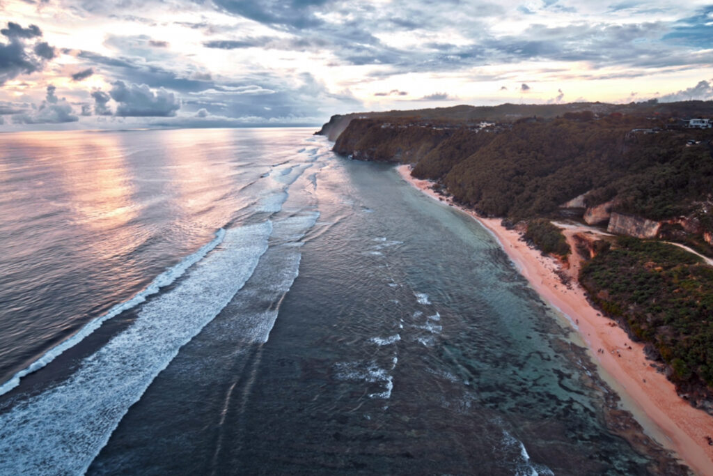 Aerial view of Bali’s dramatic coastline with turquoise water, reef breaks, and steep cliffs stretching along the shore at sunset.