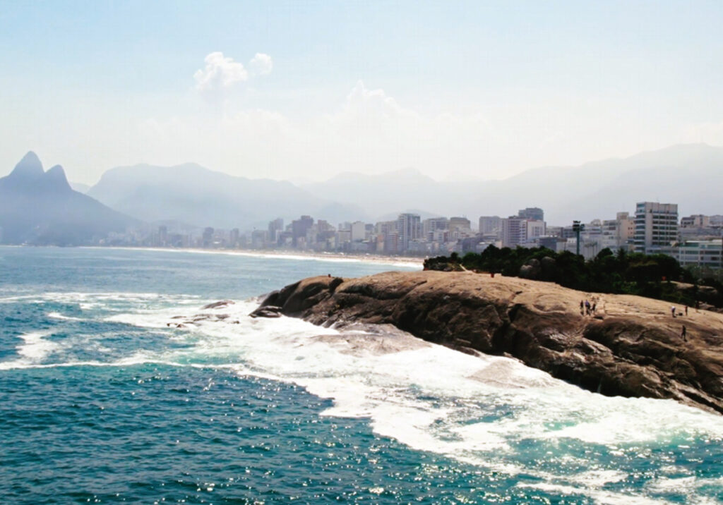 Rocky shoreline along Rio de Janeiro’s coast with waves crashing against the rocks and city buildings visible beneath distant mountains