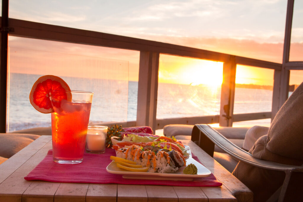 Sushi and cocktail served at a beachfront table during sunset with ocean view