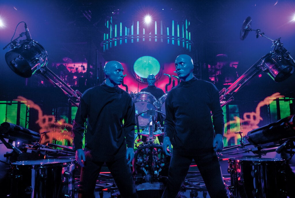 Blue Man performers on stage surrounded by percussion instruments and neon lighting during Blue Man Group in Las Vegas.