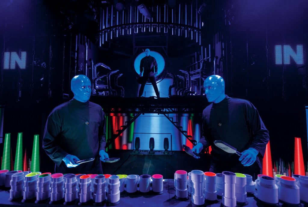 Blue Man performers playing percussion instruments surrounded by colorful stage lighting during a live Blue Man Group performance in Las Vegas