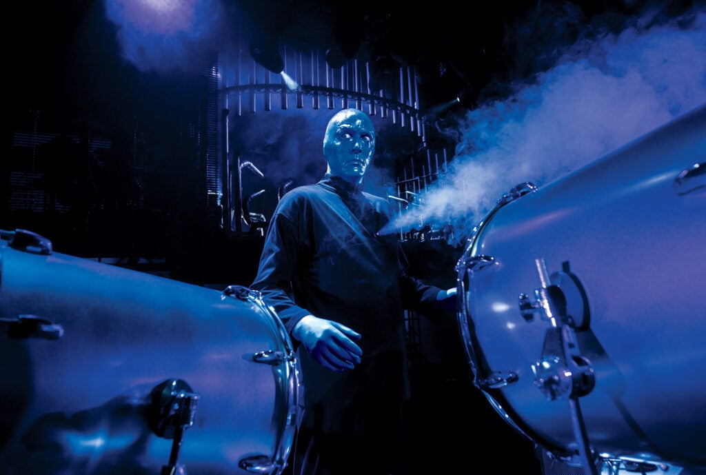 Blue Man performer surrounded by smoke and percussion instruments during a dramatic Blue Man Group performance in Las Vegas.