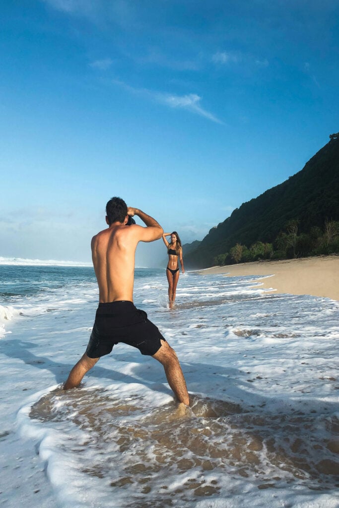 Man taking a photo of a woman in black swimwear along a tropical beach shoreline