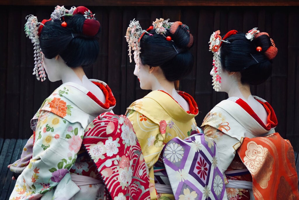 Three geisha wearing colorful traditional kimono walking together along a historic Japanese street.